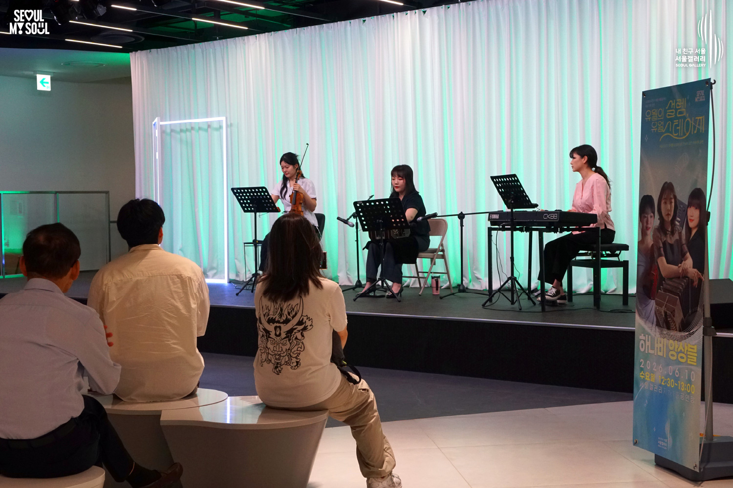 SEOUL MY SOUL 로고와 내 친구 서울 서울갤러리 SEOUL GALLERY 로고가 표기된 서울갤러리 공연장 사진으로, 푸른빛 조명이 비추는 흰색 커튼 배경의 무대 위에서 여성 연주자 세 명이 악기를 연주하며 공연을 펼치고 있으며, 무대 한쪽에는 공연 안내 배너가 설치되어 있고 객석에는 관람객들이 앉아 무대를 바라보고 있는 모습이 담긴 이미지입니다.