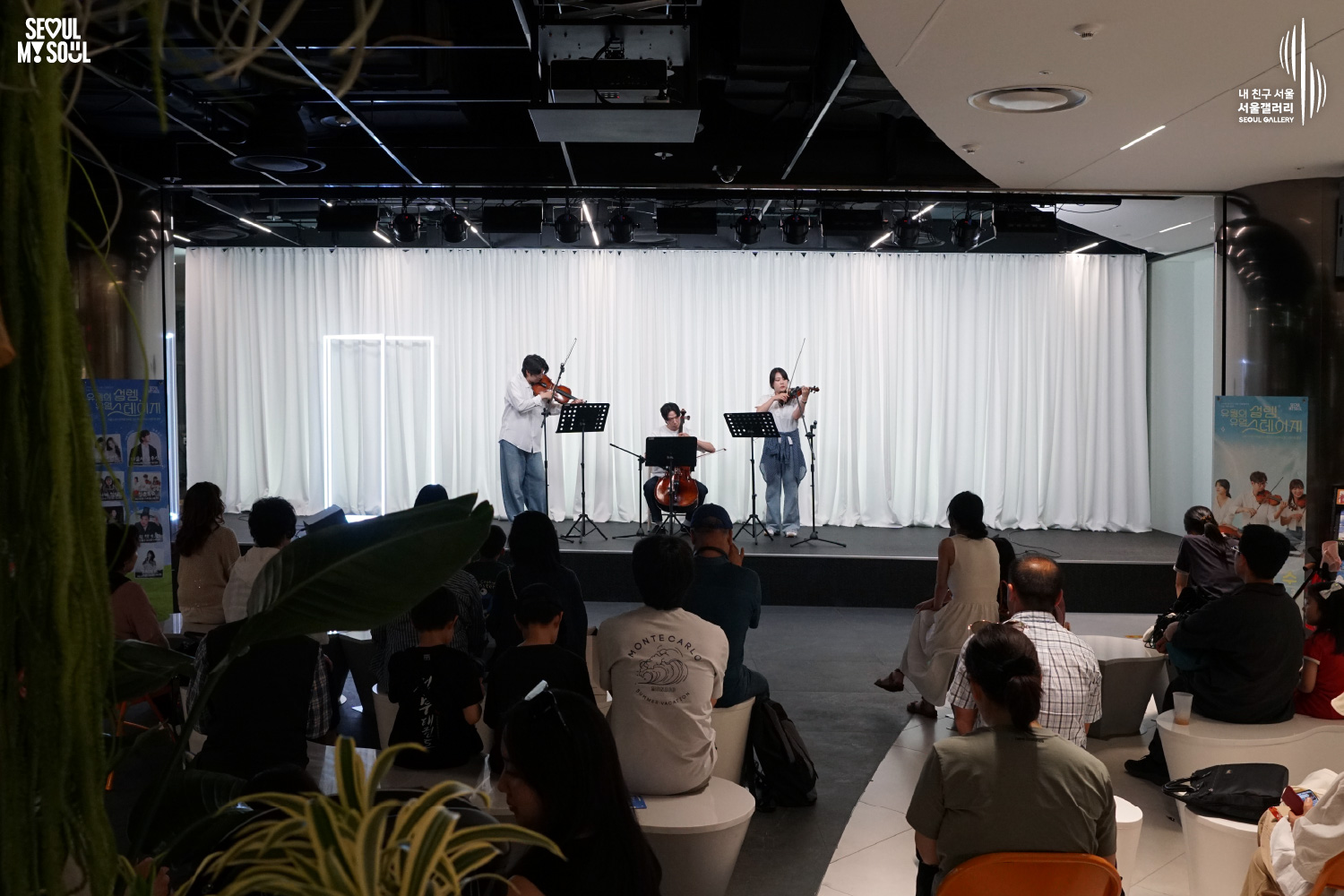 SEOUL MY SOUL 로고와 내 친구 서울 서울갤러리 SEOUL GALLERY 로고가 표기된 서울갤러리 지하 1층 공연장의 공연 현장 사진으로, 천장에 무대 조명이 설치된 실내 공간에서 흰색 커튼 배경의 무대 위에 셔츠와 바지를 입은 남성 연주자 2명과 여성 연주자 1명이 바이올린과 첼로를 연주하고 있으며, 무대 위에는 악보 거치대와 마이크 스탠드가 놓여 있고 객석에는 다양한 연령대의 관람객들이 앉아 공연을 관람하고 있는 모습이 담긴 이미지입니다.