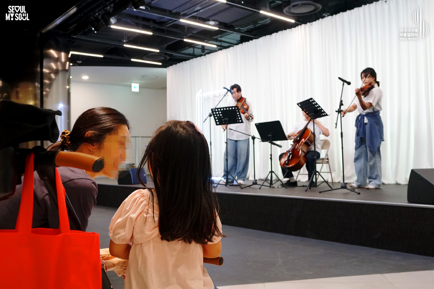 SEOUL MY SOUL 로고와 내 친구 서울 서울갤러리 SEOUL GALLERY 로고가 표기된 서울갤러리 공연장 사진으로, 천장 조명이 비추는 흰색 커튼 배경의 무대 위에서 남성 연주자 2명과 여성 연주자 1명이 바이올린과 첼로를 연주하고 있으며, 무대 앞 전경에는 관람객 두 명의 뒷모습이 보이고 객석과 무대가 함께 담긴 구도의 공연 모습이 담긴 이미지입니다.