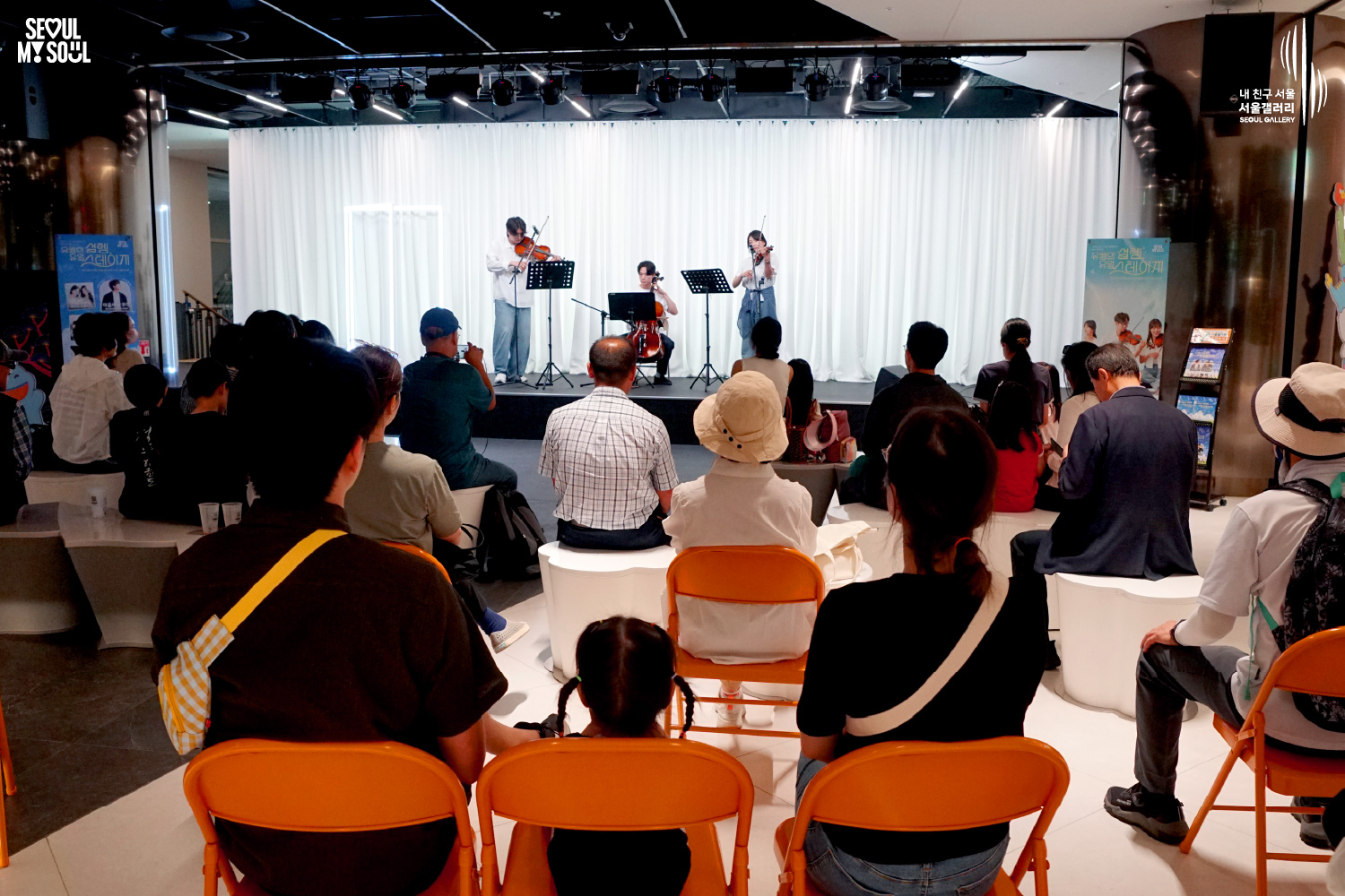SEOUL MY SOUL 로고와 내 친구 서울 서울갤러리 SEOUL GALLERY 로고가 표기된 서울갤러리 공연장 사진으로, 천장 조명이 비추는 흰색 커튼 배경의 무대 위에서 셔츠와 바지를 입은 남성 연주자 2명과 여성 연주자 1명이 바이올린과 첼로를 연주하고 있으며, 무대 양쪽에는 공연 안내판이 설치되어 있고 객석에는 다양한 연령대의 관람객들이 앉아 공연을 관람하고 있는 모습이 담긴 이미지입니다.