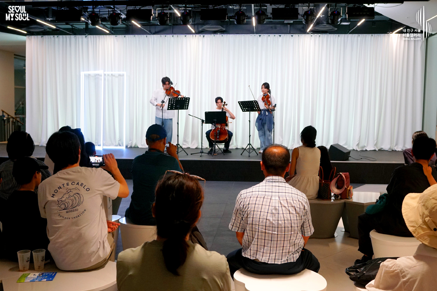 SEOUL MY SOUL 로고와 내 친구 서울 서울갤러리 SEOUL GALLERY 로고가 표기된 서울갤러리 공연장 사진으로, 천장 조명이 비추는 흰색 커튼 배경의 무대 위에서 긴 머리의 여성 연주자와 셔츠를 입은 남성 연주자들이 바이올린과 첼로를 연주하며 집중하고 있으며, 무대 위에는 악보 거치대와 마이크 스탠드가 놓여 있고 일부 관람객은 휴대전화로 무대를 촬영하고 있는 모습이 담긴 이미지입니다.