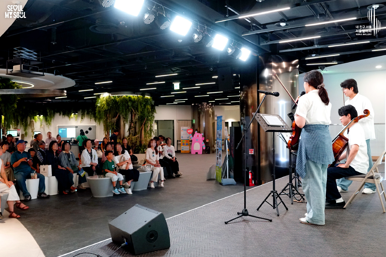 SEOUL MY SOUL 로고와 내 친구 서울 서울갤러리 SEOUL GALLERY 로고가 표기된 서울갤러리 공연장 사진으로, 천장에 설치된 무대 조명이 비추는 실내 공간에서 긴 머리의 여성 연주자가 바이올린을 들고 마이크 앞에 서 있으며, 옆에는 남성 연주자들이 바이올린과 첼로를 연주하고 있고 무대 위에는 악보 거치대와 모니터 스피커가 놓여 있으며, 무대 뒤편에서 객석 방향을 바라본 구도와 관람객들이 공연을 관람하는 모습이 담긴 이미지입니다.