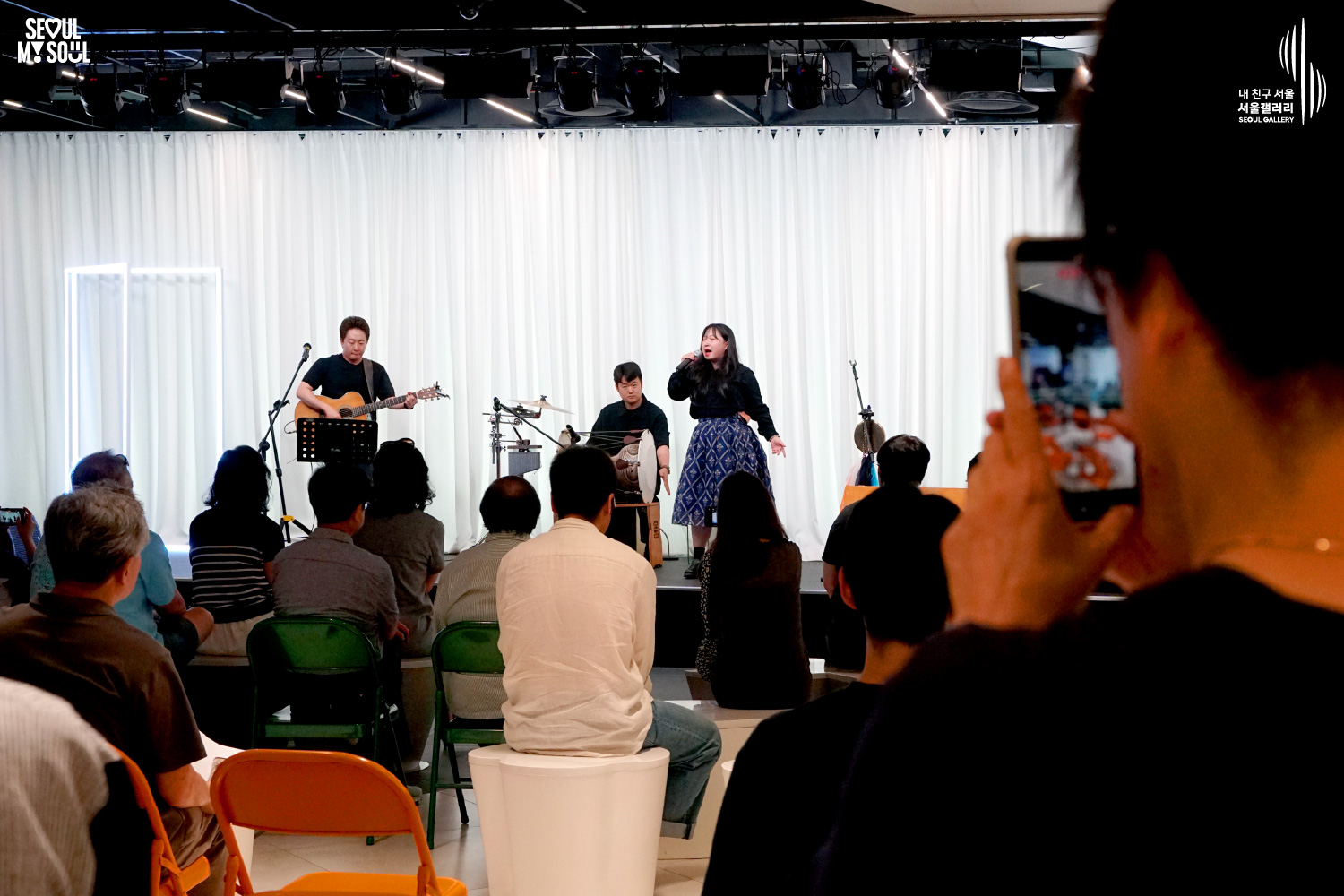 SEOUL MY SOUL 로고와 내 친구 서울 서울갤러리 SEOUL GALLERY 로고가 표기된 서울갤러리 공연장 사진으로, 천장 조명이 비추는 흰색 커튼 배경의 무대 위에서 여성 싱어송라이터가 마이크를 들고 노래를 부르고 있으며 기타를 멘 남성 싱어송라이터와 타악기 연주자가 함께 공연을 펼치고 있고, 객석에는 다양한 연령대의 관람객들이 앉아 공연을 관람하고 있으며 일부 관람객은 휴대전화로 무대를 촬영하고 있는 모습이 담긴 이미지입니다.