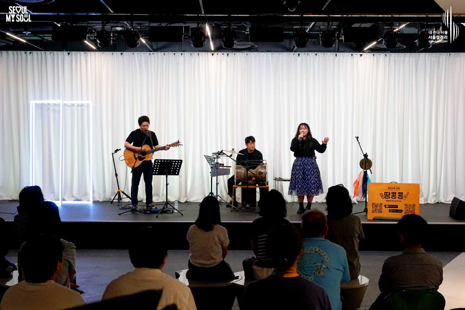 SEOUL MY SOUL 로고와 내 친구 서울 서울갤러리 SEOUL GALLERY 로고가 표기된 서울갤러리 공연장 사진으로, 사각 조명 구조물과 흰색 커튼 배경의 무대 위에서 기타를 멘 남성 싱어송라이터가 연주와 노래를 하고 있으며, 여성 싱어송라이터는 손에 든 마이크로 노래를 부르고 있고 남성 연주자는 타악기를 연주하고 있으며, 무대 위에는 마이크 스탠드와 악보 거치대, 타악기, 공연 안내판이 놓여 있고 객석에는 관람객들이 공연을 관람하고 있는 모습이 담긴 이미지입니다.