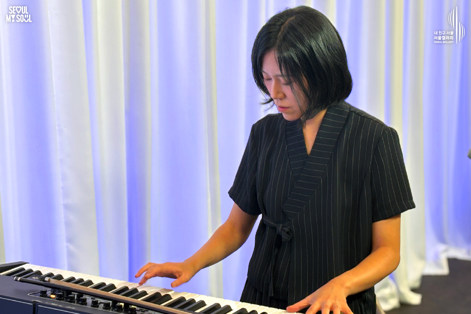 A stage photo from Seoul Gallery featuring the SEOUL MY SOUL logo and the “My Friend Seoul, Seoul Gallery” SEOUL GALLERY logo. On a stage set against a white curtain backdrop illuminated by blue lighting, the artist “Big Violin Player,” with short hair and dressed in a black top and pants, is standing and seriously playing a keyboard positioned in front of them with both hands, as shown in the image.