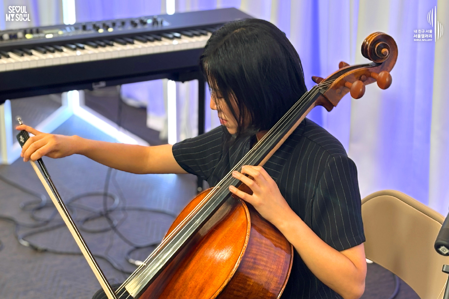 A stage photo from Seoul Gallery featuring the SEOUL MY SOUL logo and the “My Friend Seoul, Seoul Gallery” SEOUL GALLERY logo. On a stage set against a white curtain backdrop illuminated by blue lighting, the artist “Big Violin Player,” with short hair and dressed in a black top and pants, is seated on a chair, holding a cello bow in the right hand and the cello with the left hand while performing with concentration. A keyboard is positioned behind the artist, as shown in the image.