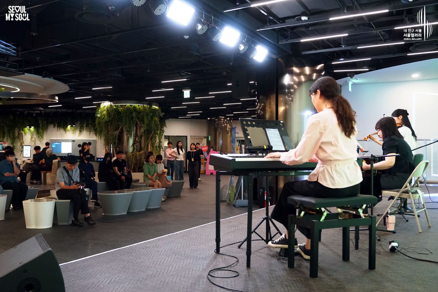 A photo of a live performance at the Seoul Gallery basement-level performance venue featuring the SEOUL MY SOUL logo and the “My Friend Seoul, Seoul Gallery” SEOUL GALLERY logo. In an indoor space equipped with overhead stage lighting, a long-haired female musician wearing a blouse is playing a keyboard, while beside her other female musicians are performing on the violin and bandoneon. Audience members of various ages are seated and watching the performance.