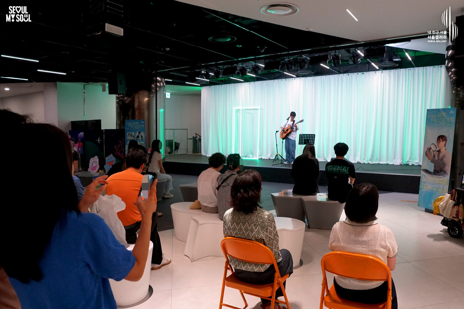 SEOUL MY SOUL 로고와 내 친구 서울 서울갤러리 SEOUL GALLERY 로고가 표기된 서울갤러리 공연장 사진으로, 연둣빛 조명과 사각 네온 조명이 어우러진 흰색 커튼 배경의 무대 위에서 안경을 착용한 남성 싱어송라이터가 어쿠스틱 기타를 연주하며 마이크 앞에서 노래를 부르고 있으며, 무대 위에는 악보 거치대와 마이크 스탠드, 공연 안내판이 놓여 있고 객석에는 다양한 연령대의 관람객들이 공연을 관람하며 일부는 휴대전화로 무대를 촬영하고 있는 모습이 담긴 이미지입니다.