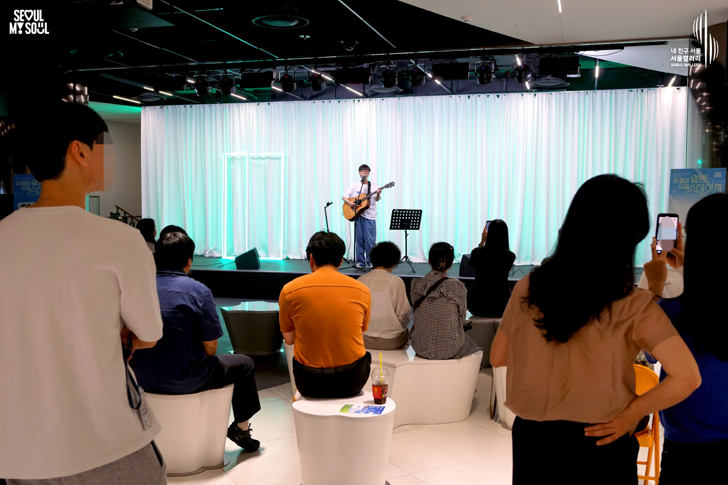 SEOUL MY SOUL 로고와 내 친구 서울 서울갤러리 SEOUL GALLERY 로고가 표기된 서울갤러리 공연장 사진으로, 연둣빛 조명과 사각 네온 조명이 어우러진 흰색 커튼 배경의 무대 위에서 안경을 착용한 남성 싱어송라이터가 어쿠스틱 기타를 연주하며 마이크 앞에서 노래를 부르고 있으며, 무대 위에는 악보 거치대와 마이크 스탠드, 모니터 스피커가 놓여 있고 객석에는 관람객들이 공연을 관람하며 일부는 휴대전화로 무대를 촬영하고 있는 모습이 담긴 이미지입니다.