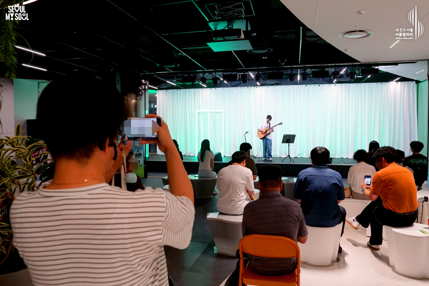 SEOUL MY SOUL 로고와 내 친구 서울 서울갤러리 SEOUL GALLERY 로고가 표기된 서울갤러리 공연장 사진으로, 연둣빛 조명과 사각 네온 조명이 어우러진 흰색 커튼 배경의 무대 위에서 안경을 착용한 남성 싱어송라이터가 어쿠스틱 기타를 연주하며 노래를 부르고 있으며, 무대 위에는 마이크 스탠드와 악보 거치대가 놓여 있고 객석에는 다양한 연령대의 관람객들이 공연을 관람하며 전경의 관람객이 휴대전화로 무대를 촬영하고 있는 모습이 담긴 이미지입니다.