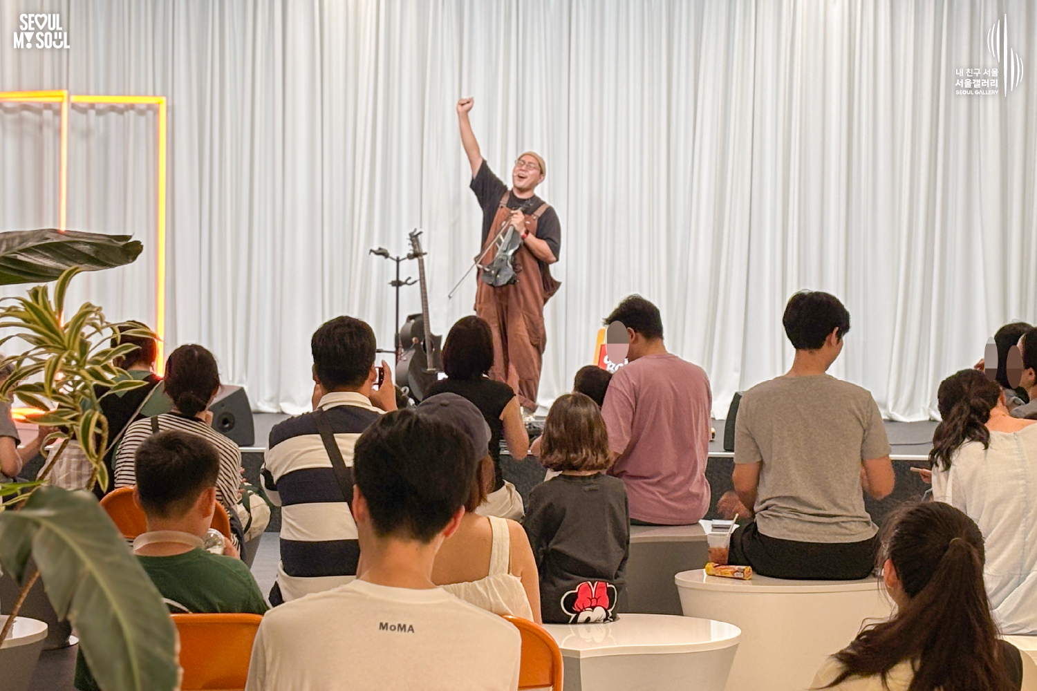 SEOUL MY SOUL 로고와 내 친구 서울 서울갤러리 SEOUL GALLERY 로고가 표기된 서울갤러리 공연장 사진으로, 흰색 커튼 배경의 무대 위에서 비니 모자와 안경을 착용한 멜빵 의상의 남성 연주자가 한 손을 들어 관객에게 인사하고 있으며, 무대 위에는 기타 거치대와 마이크 스탠드, 모니터 스피커가 놓여 있고 객석에는 다양한 연령대의 관람객들이 공연을 관람하며 일부는 휴대전화로 무대를 촬영하고 있는 모습이 담긴 이미지입니다.