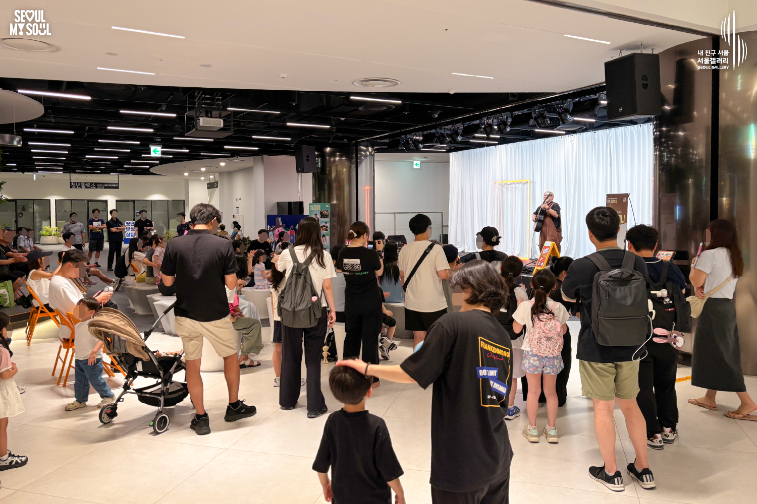 SEOUL MY SOUL 로고와 내 친구 서울 서울갤러리 SEOUL GALLERY 로고가 표기된 서울갤러리 지하 1층 공연장의 공연 현장 사진으로, 천장에 무대 조명이 설치된 실내 공간에서 비니 모자와 안경을 착용한 멜빵 의상의 남성 연주자가 기타를 연주하고 있으며, 무대 위에는 마이크 스탠드와 공연 안내판이 놓여 있고 객석에는 어린이를 포함한 다양한 연령대의 관람객들이 공연을 관람하며 일부는 서서 무대를 바라보고 있는 모습이 담긴 이미지입니다.