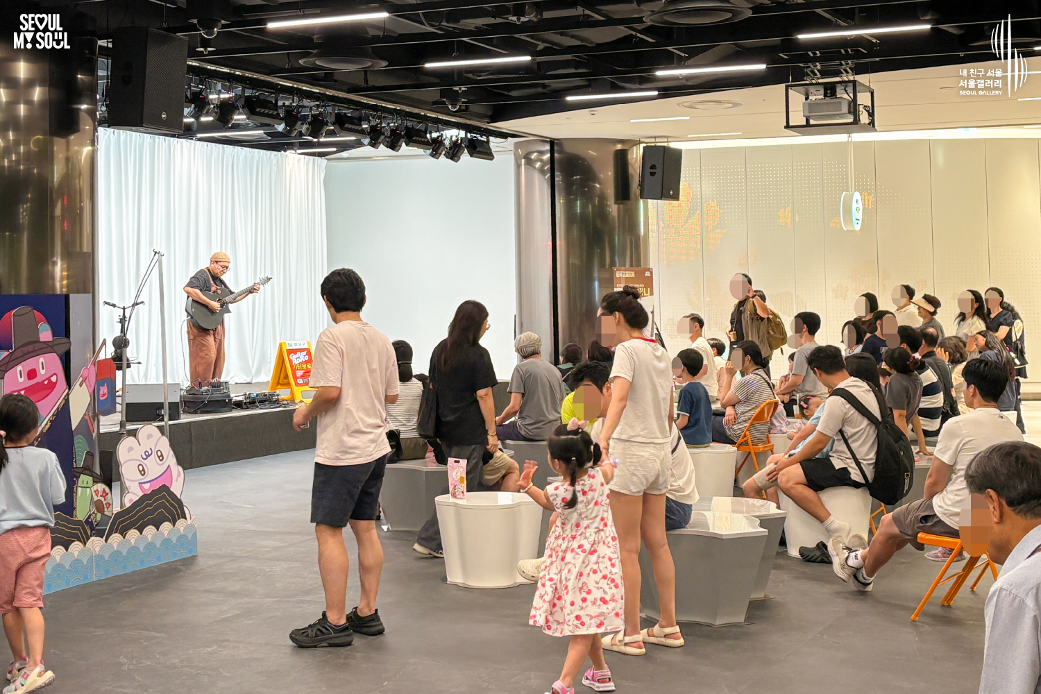 SEOUL MY SOUL 로고와 내 친구 서울 서울갤러리 SEOUL GALLERY 로고가 표기된 서울갤러리 공연장 사진으로, 천장에 무대 조명이 설치된 실내 공간에서 비니 모자와 안경을 착용한 멜빵 의상의 남성 연주자가 기타를 연주하고 있으며, 무대 위에는 마이크 스탠드와 공연 안내판이 놓여 있고 객석에는 어린이를 포함한 다양한 연령대의 관람객들이 공연을 관람하고 있는 모습이 담긴 이미지입니다.