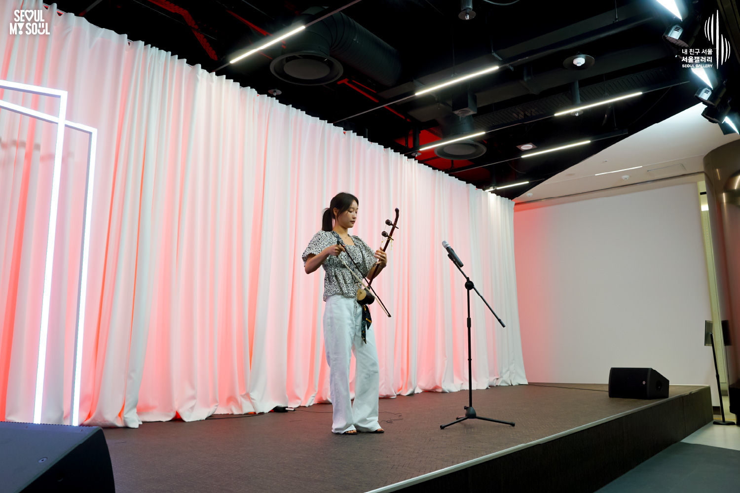 SEOUL MY SOUL 로고와 내 친구 서울 서울갤러리 SEOUL GALLERY 로고가 표기된 서울갤러리 공연장 무대 사진으로, 분홍빛 조명과 사각 네온 조명이 어우러진 흰색 커튼 배경의 무대 위에서 긴 머리를 묶은 여성 연주자 우하린이 해금을 연주하고 있으며, 무대 위에는 마이크 스탠드와 모니터 스피커가 놓여 있는 모습이 담긴 이미지입니다.