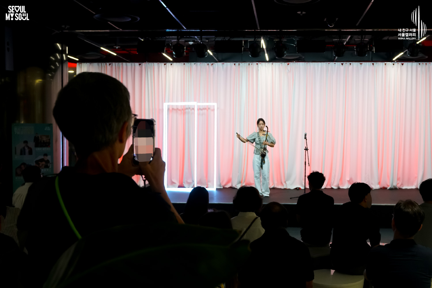SEOUL MY SOUL 로고와 내 친구 서울 서울갤러리 SEOUL GALLERY 로고가 표기된 서울갤러리 공연장 사진으로, 분홍빛 조명과 사각 네온 조명이 어우러진 흰색 커튼 배경의 무대 위에서 긴 머리를 묶은 여성 연주자 우하린이 해금을 연주하고 있으며, 무대 위에는 마이크 스탠드가 놓여 있고 객석에는 다양한 연령대의 관람객들이 공연을 관람하고 있으며 일부 관람객은 휴대전화로 무대를 촬영하고 있는 모습이 담긴 이미지입니다.