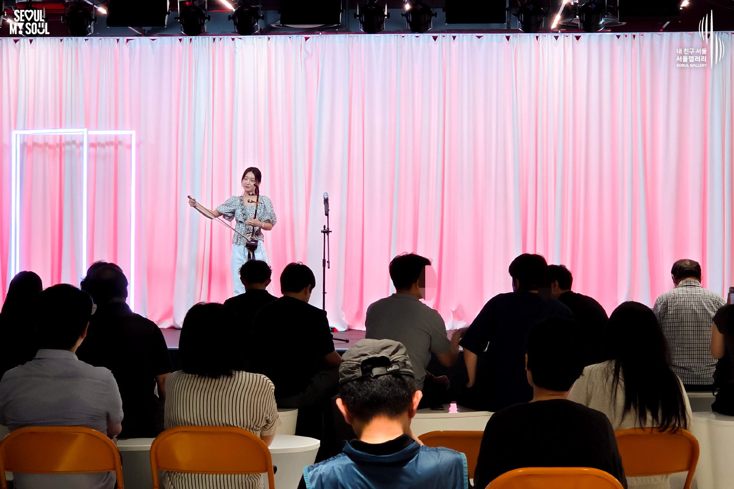 SEOUL MY SOUL 로고와 내 친구 서울 서울갤러리 SEOUL GALLERY 로고가 표기된 서울갤러리 공연장 사진으로, 분홍빛 조명이 비추는 흰색 커튼 배경의 무대 위에서 긴 머리를 묶은 여성 연주자 우하린이 해금을 연주하고 있으며, 무대 위에는 마이크 스탠드가 놓여 있고 객석에는 다양한 연령대의 관람객들이 공연을 관람하고 있는 모습이 담긴 이미지입니다.
