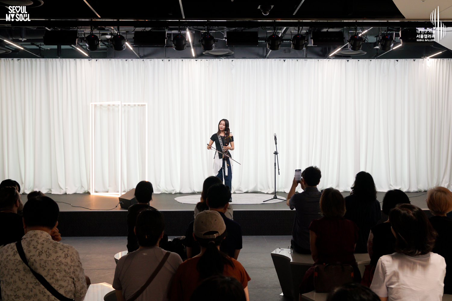 SEOUL MY SOUL 로고와 내 친구 서울 서울갤러리 SEOUL GALLERY 로고가 표기된 서울갤러리 공연장 사진으로, 천장 무대 조명이 비추는 흰색 커튼 배경의 무대 위에서 긴 머리에 상의와 바지를 입은 여성 연주자가 해금을 들고 활을 잡고 서 있으며, 무대에는 마이크 스탠드와 스피커가 놓여 있고 객석에는 관람객들이 앉아 무대를 바라보며 일부는 휴대전화로 촬영하고 있는 모습이 담긴 이미지입니다.