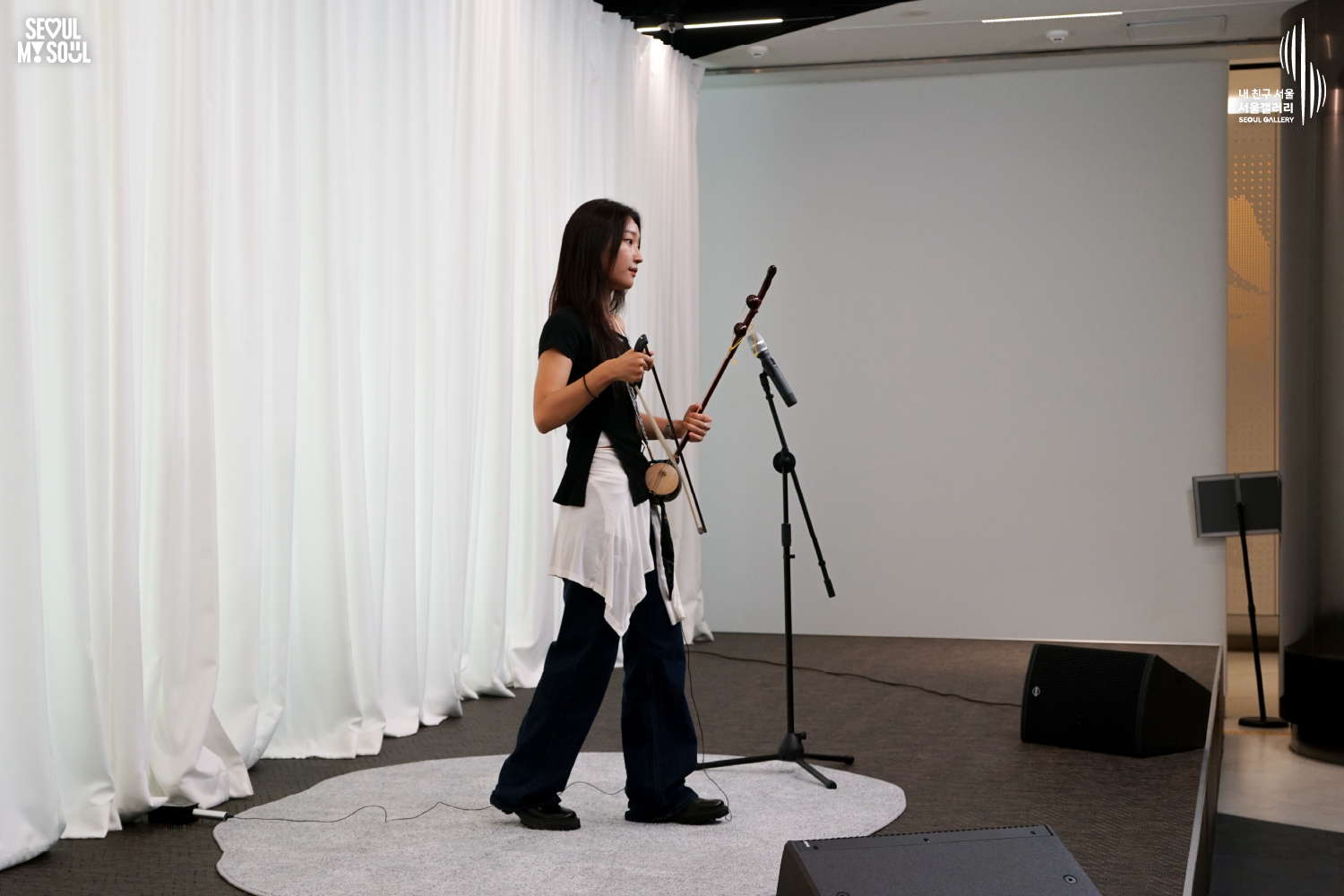 SEOUL MY SOUL 로고와 내 친구 서울 서울갤러리 SEOUL GALLERY 로고가 표기된 서울갤러리 공연장 무대 사진으로, 천장 조명이 비추는 흰색 커튼 배경의 무대 위에서 긴 머리에 상의와 바지를 입은 여성 연주자가 해금을 몸 앞에 들고 한 손에 활을 잡은 채 마이크 앞에 서 있으며, 주변에는 마이크 스탠드와 모니터 스피커가 놓여 있는 모습이 담긴 이미지입니다.