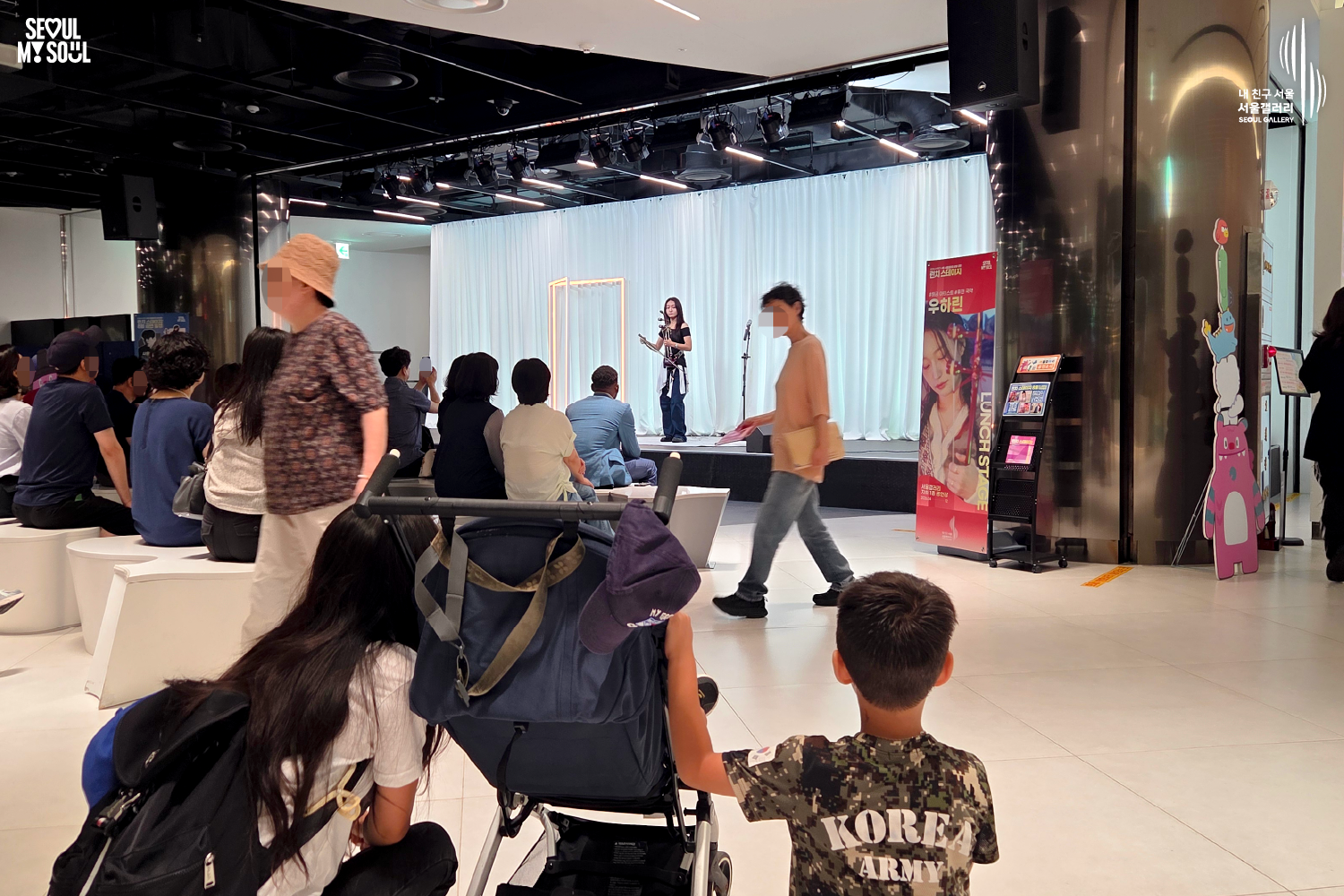 SEOUL MY SOUL 로고와 내 친구 서울 서울갤러리 SEOUL GALLERY 로고가 표기된 서울갤러리 지하 1층 공연장의 공연 현장 사진으로, 천장 무대 조명이 비추는 흰색 커튼 배경의 무대 위에서 긴 머리의 여성 연주자가 해금을 들고 공연하고 있으며, 무대 앞에는 관람객들이 앉아 있고 전경에는 유모차와 어린이, 공연장 앞을 지나가는 관람객들이 함께 보이는 모습이 담긴 이미지입니다.