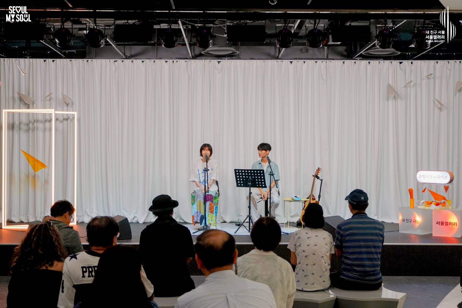 SEOUL MY SOUL 로고와 내 친구 서울 서울갤러리 SEOUL GALLERY 로고가 표기된 서울갤러리 공연장 사진으로, 천장 무대 조명과 사각 조명이 비추고 종이비행기 장식이 달린 흰색 커튼 배경의 무대에서 짧은 머리의 여성 아티스트와 남성 아티스트가 의자에 앉아 마이크 앞에서 공연하고 있으며, 무대에는 악보대와 기타, 스피커, 종이 조형물이 놓여 있고 객석에는 관람객들이 앉아 무대를 바라보는 모습이 담긴 이미지입니다.