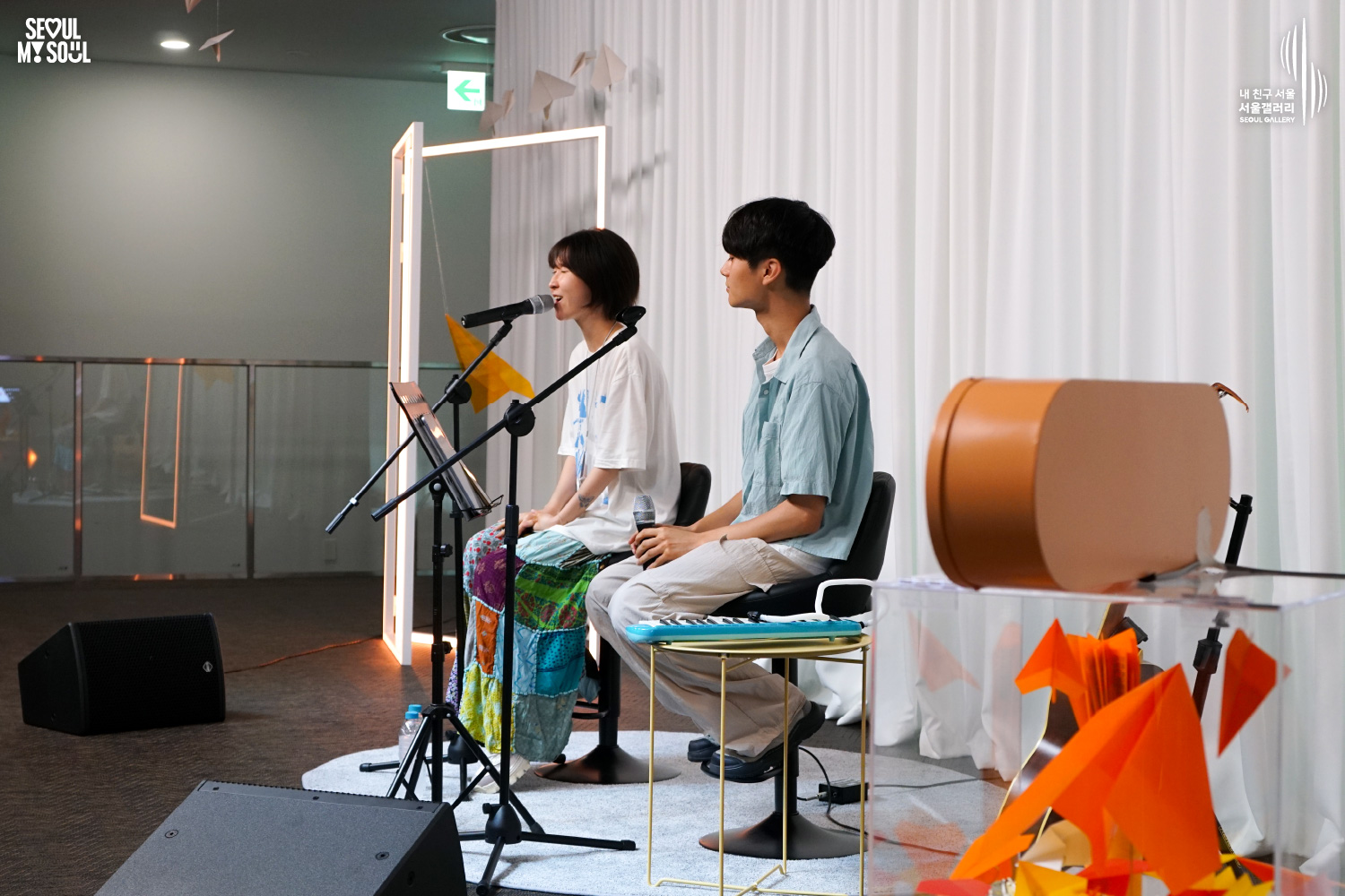 SEOUL MY SOUL 로고와 내 친구 서울 서울갤러리 SEOUL GALLERY 로고가 표기된 서울갤러리 공연장 무대 사진으로, 천장 조명과 사각 조명이 비추는 흰색 커튼 배경의 무대에서 짧은 머리의 여성 아티스트가 의자에 앉아 마이크 앞에서 노래하고 있으며, 옆에는 짧은 머리의 남성 아티스트가 손에 마이크를 든 채 앉아 있고 주변에는 마이크 스탠드와 건반악기, 종이비행기 장식이 놓여 있는 모습이 담긴 이미지입니다.