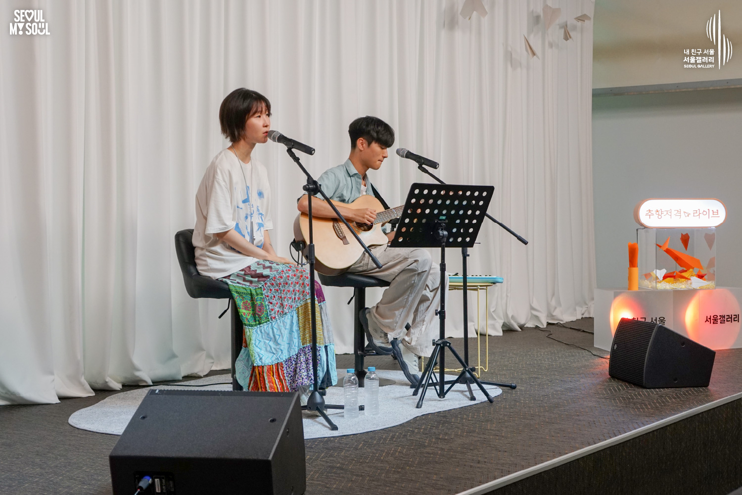 SEOUL MY SOUL 로고와 내 친구 서울 서울갤러리 SEOUL GALLERY 로고가 표기된 서울갤러리 공연장 무대 사진으로, 천장 조명이 비추고 종이비행기 장식이 달린 흰색 커튼 배경의 무대에서 짧은 머리의 여성 아티스트가 의자에 앉아 마이크 앞에서 노래하고 있으며, 옆에서는 짧은 머리의 남성 아티스트가 의자에 앉아 어쿠스틱 기타를 연주하고 있고 앞에는 악보대와 모니터 스피커가 놓여 있는 모습이 담긴 이미지입니다.