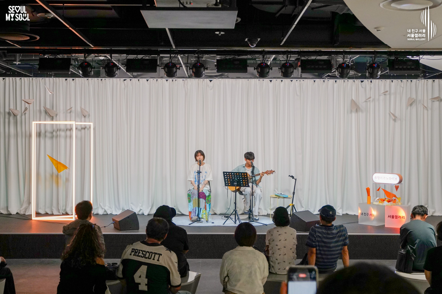 SEOUL MY SOUL 로고와 내 친구 서울 서울갤러리 SEOUL GALLERY 로고가 표기된 서울갤러리 공연장 사진으로, 천장 무대 조명과 사각 조명이 비추고 종이비행기 장식이 달린 흰색 커튼 배경의 무대에서 짧은 머리의 여성 아티스트가 마이크 앞에서 노래하고 짧은 머리의 남성 아티스트가 어쿠스틱 기타를 연주하고 있으며, 객석에는 관람객들이 앉아 공연을 바라보고 전경에는 휴대전화를 든 관람객이 보이는 모습이 담긴 이미지입니다.