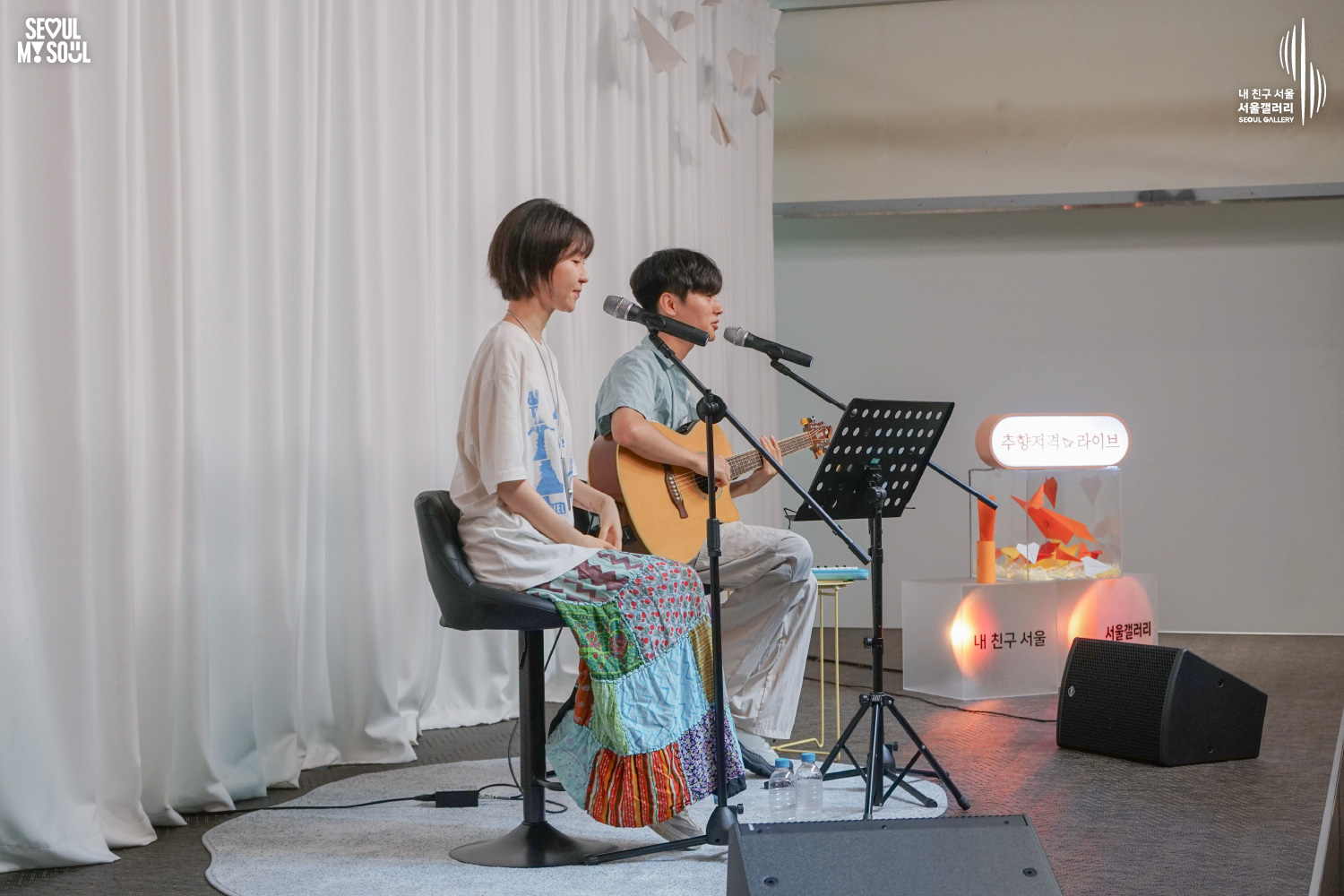 SEOUL MY SOUL 로고와 내 친구 서울 서울갤러리 SEOUL GALLERY 로고가 표기된 서울갤러리 공연장 무대 사진으로, 천장 조명이 비추고 종이비행기 장식이 달린 흰색 커튼 배경의 무대에서 짧은 머리의 여성 아티스트가 의자에 앉아 있으며, 옆의 짧은 머리 남성 아티스트가 어쿠스틱 기타를 연주하며 마이크 앞에서 노래하고 있고 앞에는 악보대와 스피커가 놓여 있는 모습이 담긴 이미지입니다.