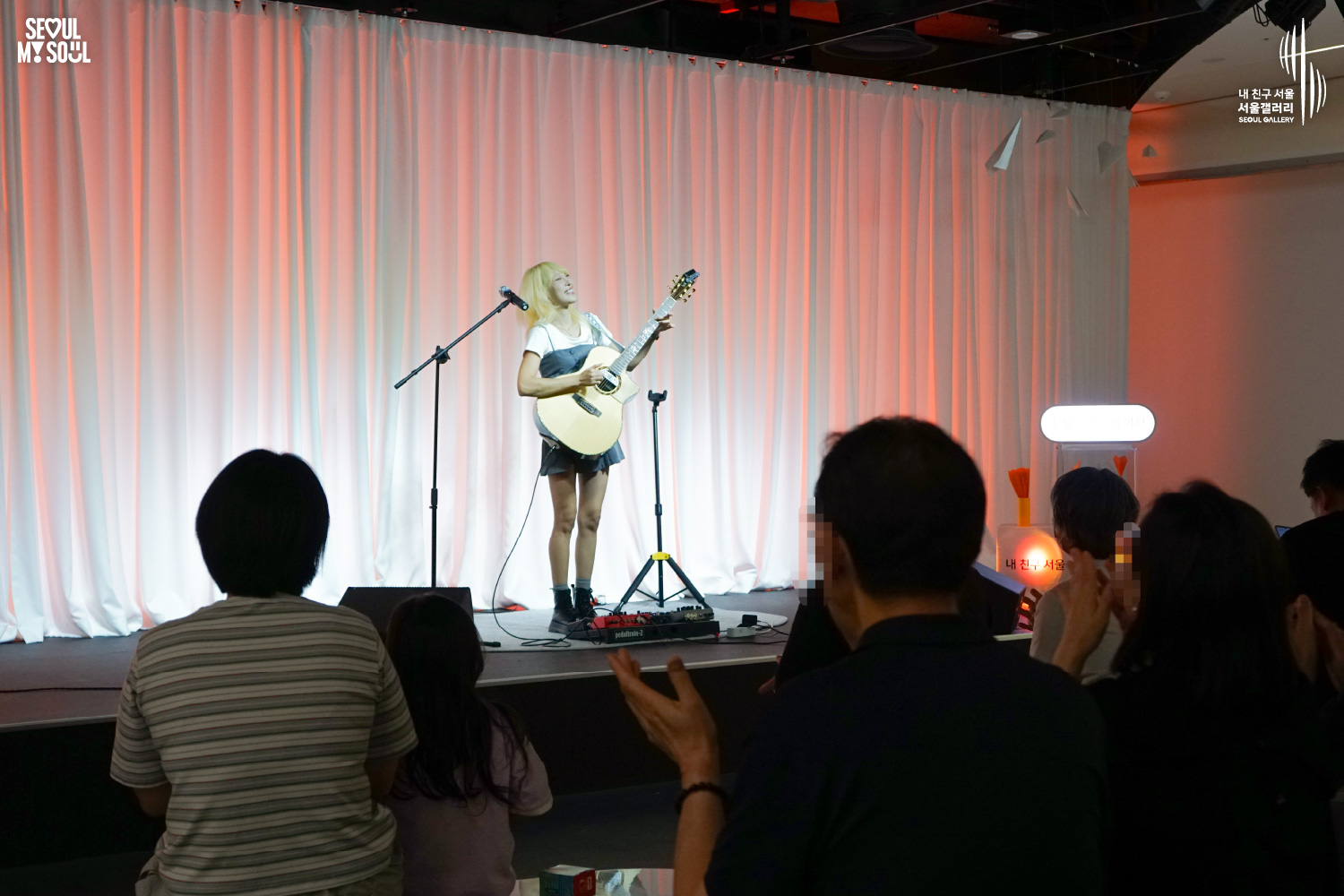 SEOUL MY SOUL 로고와 내 친구 서울 서울갤러리 SEOUL GALLERY 로고가 표기된 서울갤러리 공연장 사진으로, 주황빛 조명이 비추는 흰색 커튼 배경의 무대 위에서 긴 머리의 여성 연주자 김나린이 어쿠스틱 기타를 연주하고 있으며, 마이크 스탠드와 페달 이펙터가 놓여 있고 객석의 관람객들이 무대를 바라보거나 박수를 치는 모습이 담긴 이미지입니다.