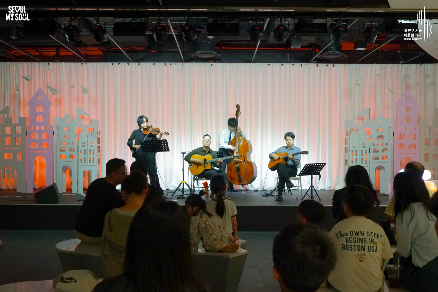 SEOUL MY SOUL 로고와 내 친구 서울 서울갤러리 SEOUL GALLERY 로고가 표기된 서울갤러리 공연장 사진으로, 주황빛 조명이 비추는 흰색 커튼과 건물 모양 장식 앞에서 라쁘띠프랑스콰르텟의 여성 바이올린 연주자와 남성 더블베이스 연주자, 두 명의 남성 기타 연주자가 함께 연주하고 있으며, 객석에는 다양한 연령대의 관람객들이 무대를 바라보는 모습이 담긴 이미지입니다.