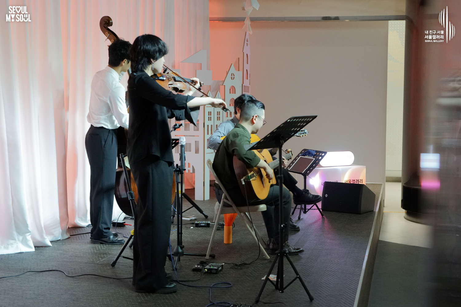 SEOUL MY SOUL 로고와 내 친구 서울 서울갤러리 SEOUL GALLERY 로고가 표기된 서울갤러리 공연장 무대 사진으로, 주황빛 조명이 비추는 흰색 커튼과 건물 모양 장식 앞에서 라쁘띠프랑스콰르텟의 여성 연주자가 활로 바이올린을 연주하고 있으며, 뒤편에는 더블베이스와 기타를 연주하는 남성 연주자들이 있고 악보대와 의자가 놓여 있는 이미지입니다.