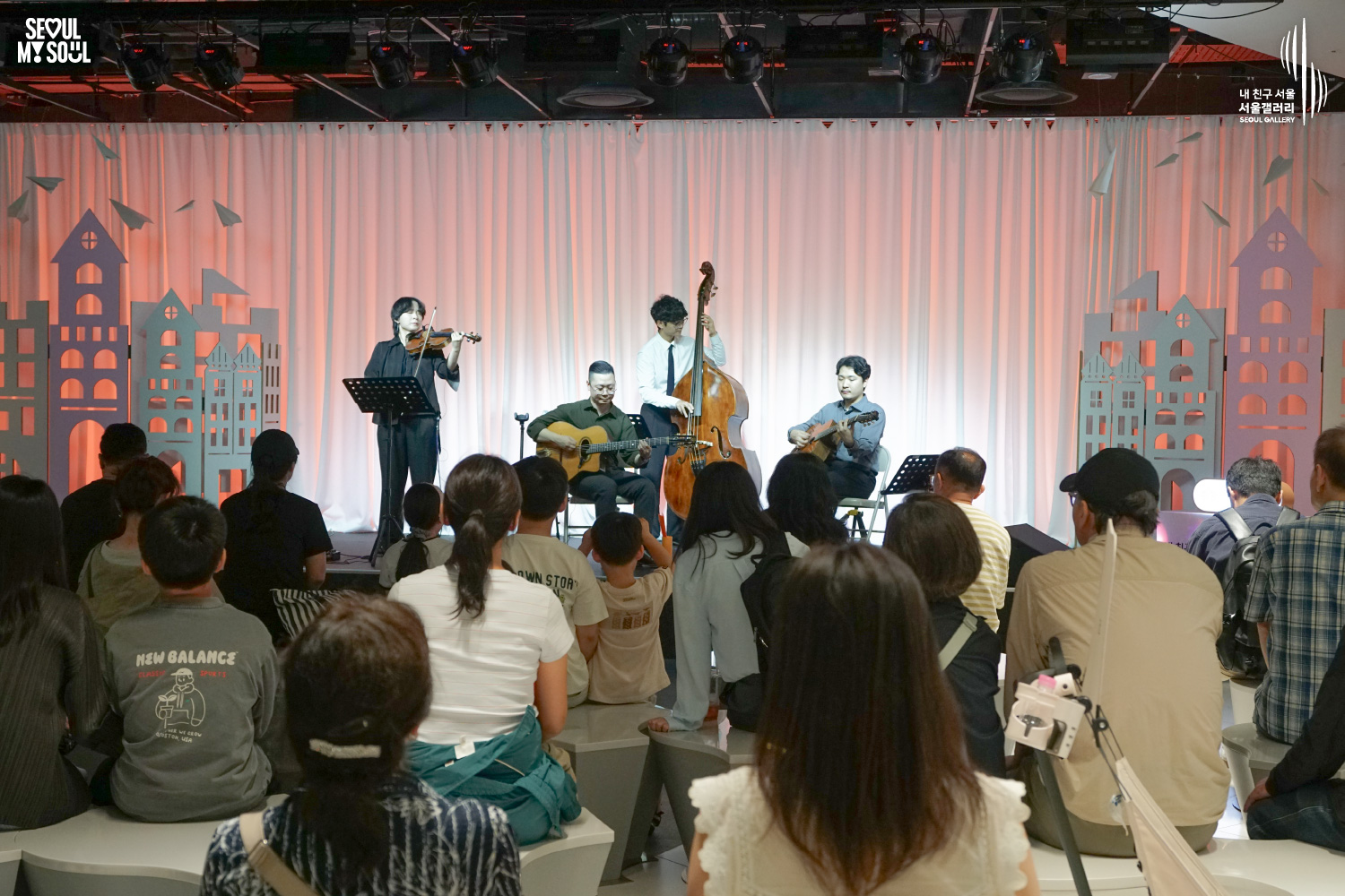 SEOUL MY SOUL 로고와 내 친구 서울 서울갤러리 SEOUL GALLERY 로고가 표기된 서울갤러리 공연장 사진으로, 주황빛 조명이 비추는 흰색 커튼 배경의 무대에서 라쁘띠프랑스콰르텟의 여성 바이올린 연주자와 남성 더블베이스 연주자, 두 명의 남성 기타 연주자가 함께 연주하고 있으며, 객석 전경에는 관람객들의 뒷모습이 가득 보이는 이미지입니다.
