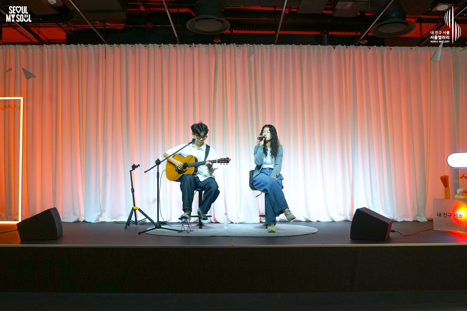 SEOUL MY SOUL 로고와 내 친구 서울 서울갤러리 SEOUL GALLERY 로고가 표기된 서울갤러리 공연장 무대 사진으로, 주황빛 조명이 비추는 흰색 커튼 배경의 무대에서 안경을 착용한 남성 연주자와 긴 곱슬머리의 여성 보컬로 구성된 아웃오브캠퍼스가 의자에 앉아 기타를 연주하고 마이크로 노래를 부르고 있으며, 마이크 스탠드와 모니터 스피커가 놓여 있는 이미지입니다.